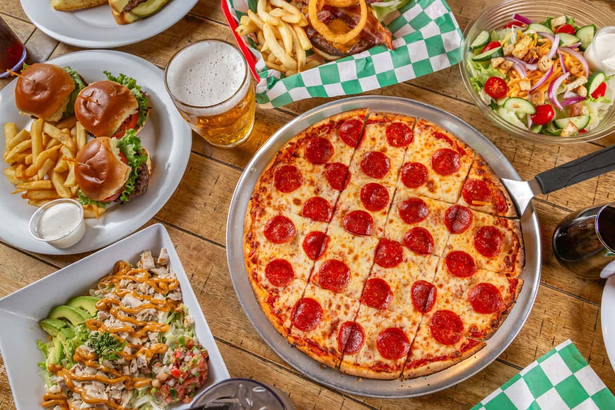 Pepperoni pizza, mini burgers, fries, salads, and drinks on a wooden table in a lively casual dining setting.