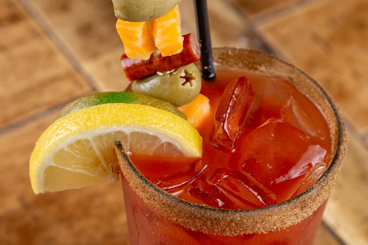 Bloody Mary garnished with lemon, lime, olives, cheese, and sausage skewer in a chili-rimmed glass with ice.