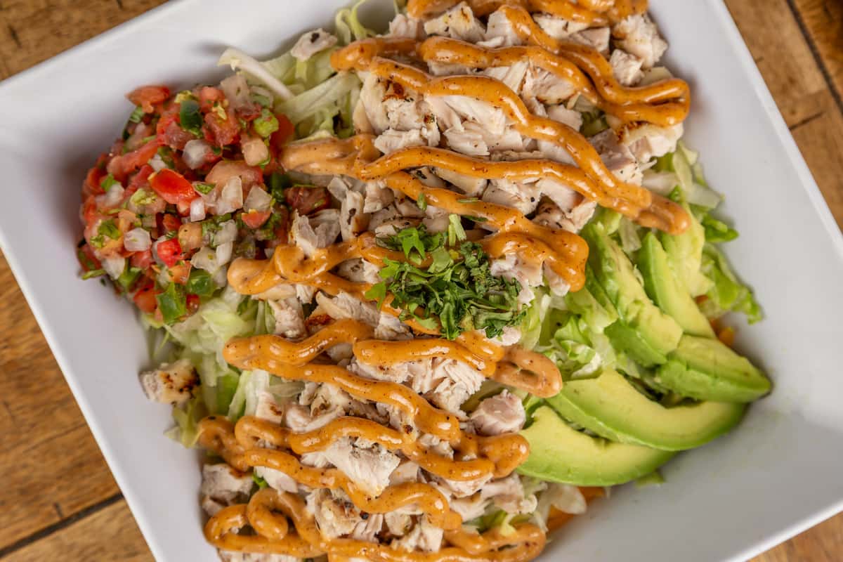 Chopped grilled chicken salad with shredded lettuce, avocado, pico de gallo, and spicy dressing in a white square bowl.