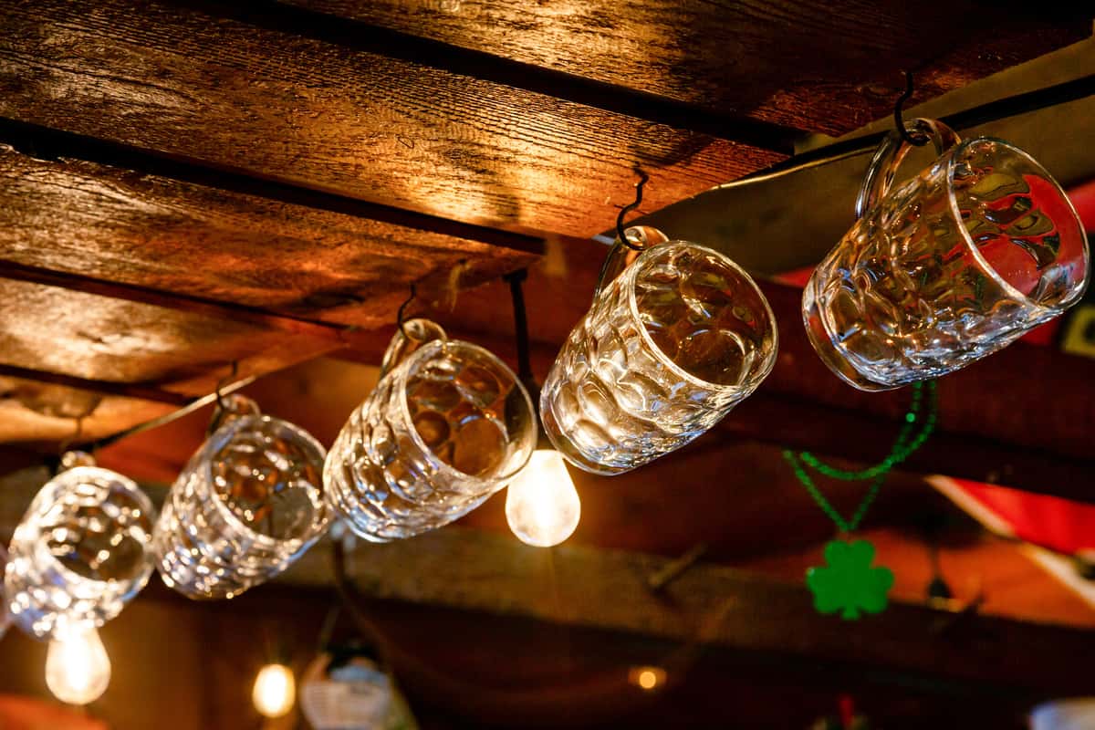 Clear glass beer mugs hanging upside down from hooks on a rustic wooden ceiling, warmly lit by exposed bulbs.