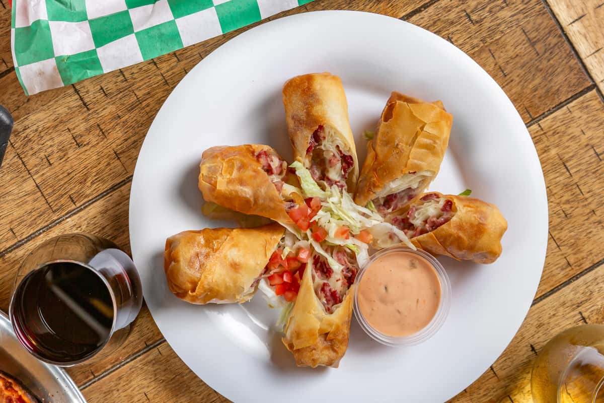 Crispy egg rolls filled with corned beef and cheese, served with shredded lettuce, diced tomatoes, and a side of dipping sauce.