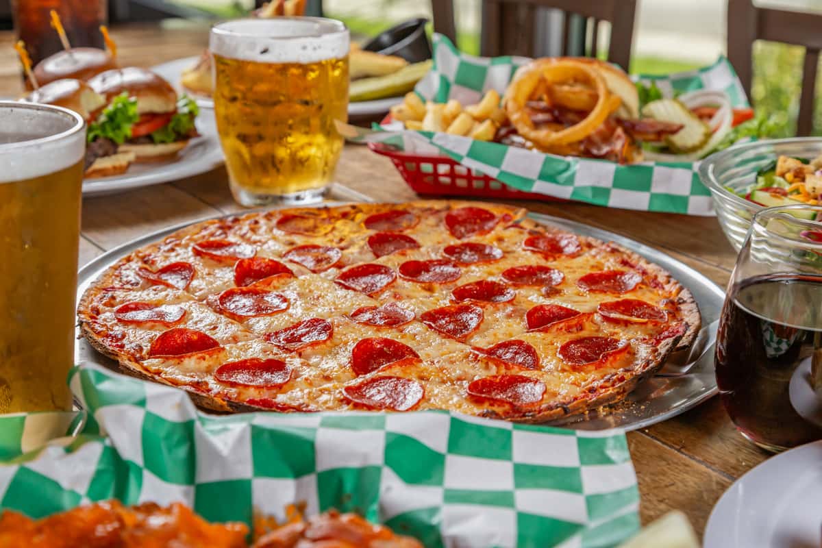 Pepperoni pizza on a tray surrounded by beer, wine, burgers, fries, wings, and salad on a rustic wooden table.
