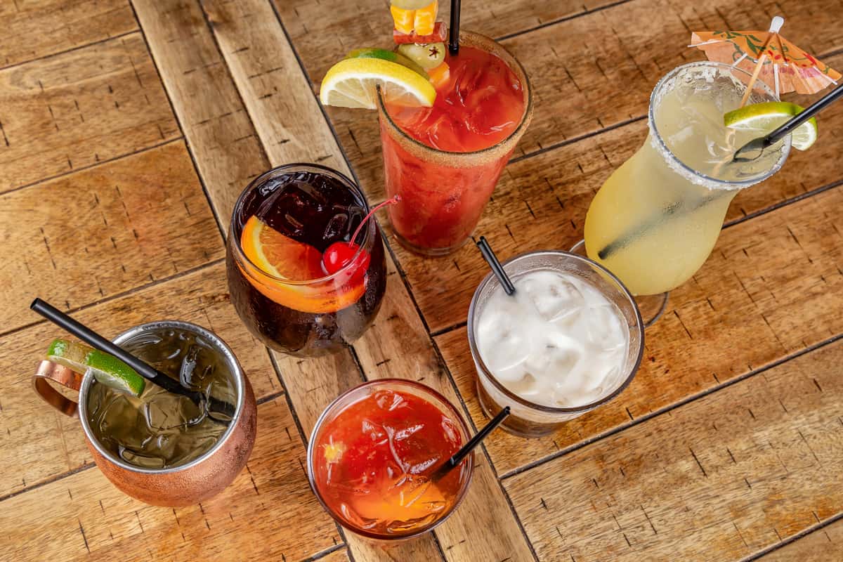 Assorted cocktails on a wooden table, including a margarita, Bloody Mary, Moscow mule, and drinks garnished with fruit.