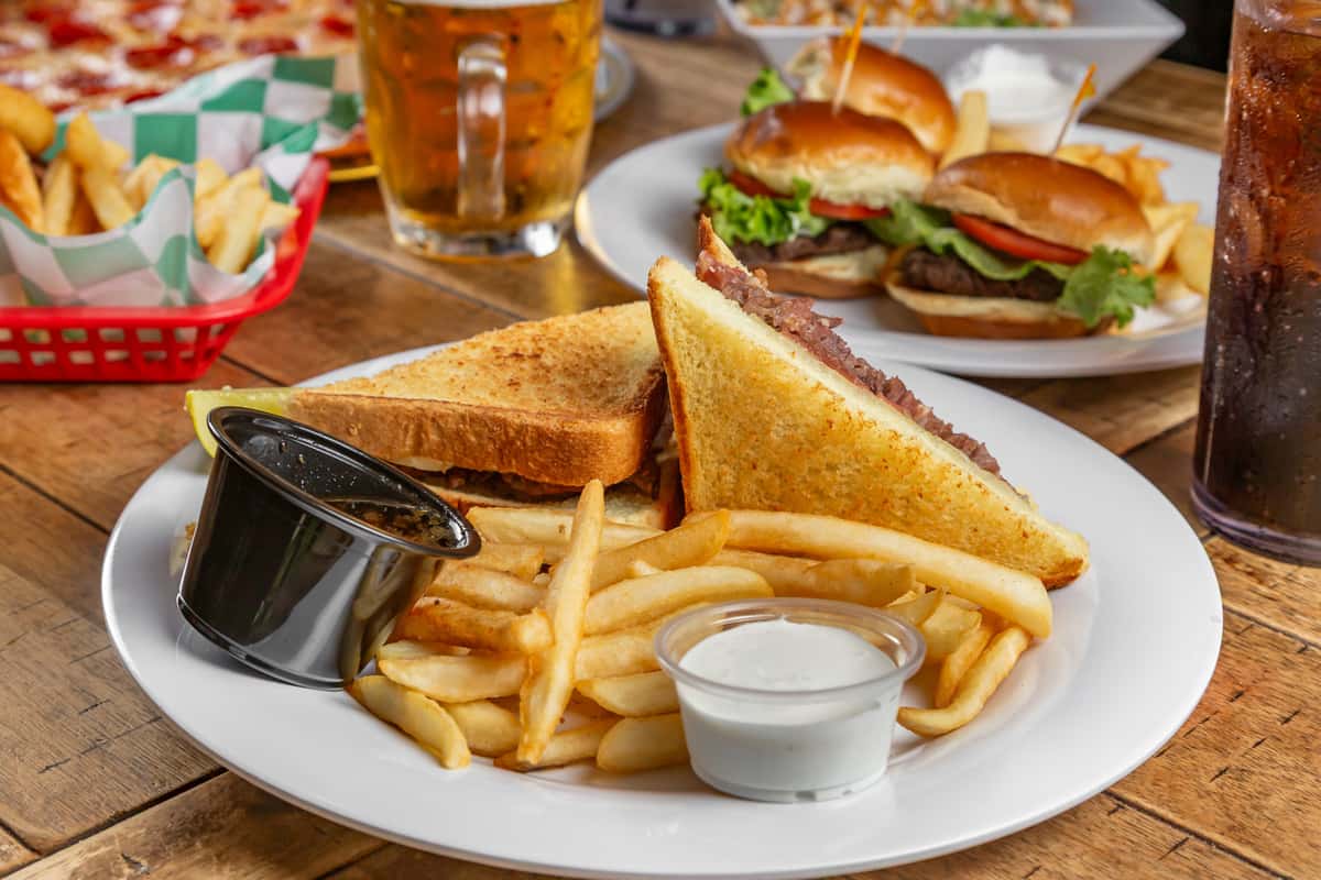 Grilled sandwich with fries, pickle, au jus, and ranch on a white plate, with burgers, beer, and soda in the background.