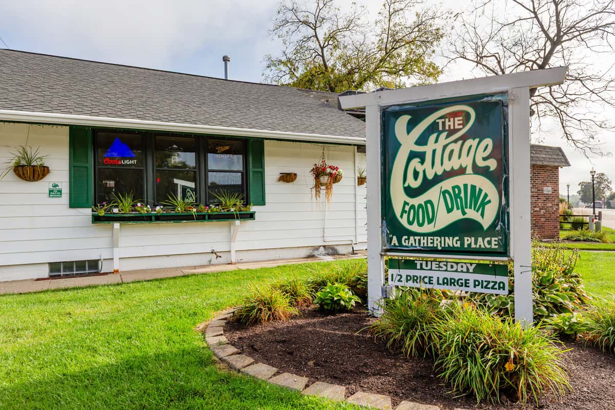 A cozy white building with green shutters and flower boxes, featuring a large sign that reads **"The Cottage Food/Drink – A Gathering Place"** and promoting **half-price large pizza on Tuesdays**. The establishment has a welcoming, homey appearance with a well-maintained lawn and visible bar signage in the window.