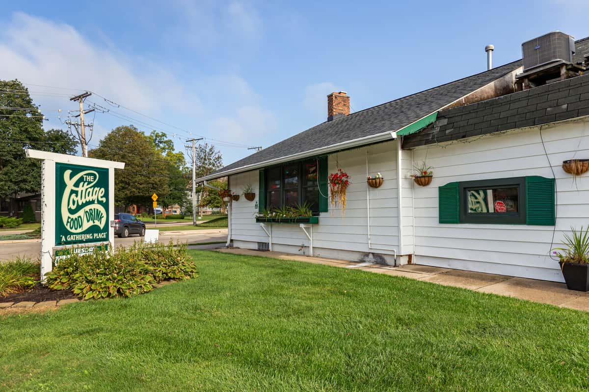 White building with green shutters and flower baskets, featuring The Cottage Food/Drink sign on a grassy corner lot.