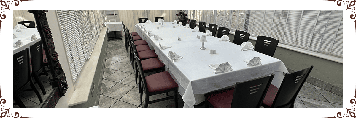 Elegant indoor dining space with long white tablecloth-covered table, neatly set plates, wine glasses, and red-cushioned chairs.
