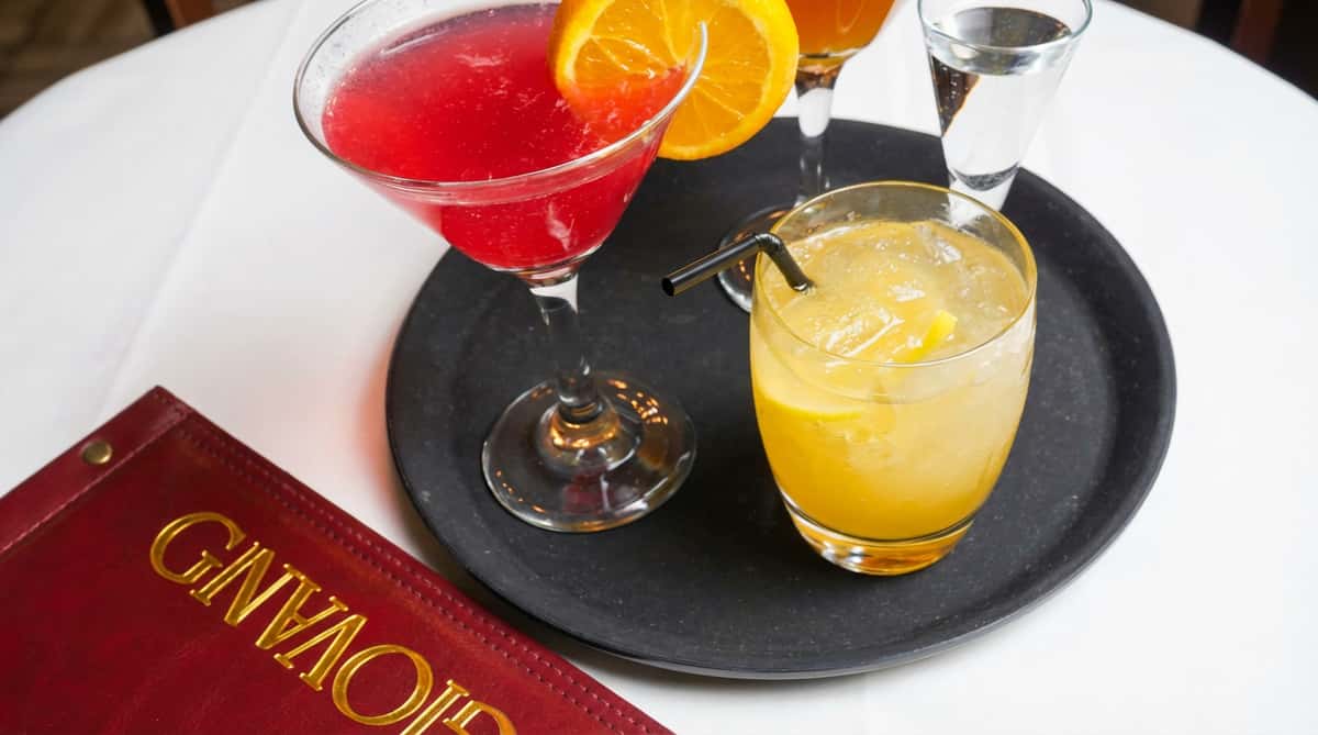 A red cocktail garnished with an orange slice, a yellow cocktail with a black straw, and a shot glass of clear liquid on a black tray.