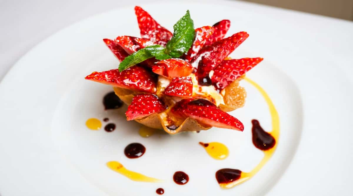 Strawberry dessert in a waffle cup with cream, powdered sugar, mint leaves, and balsamic glaze on a white plate.