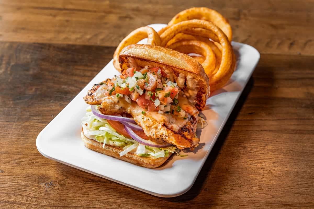 spicy crispy chicken sandwich topped with diced peppers and sauce served with onion rings on a white platter