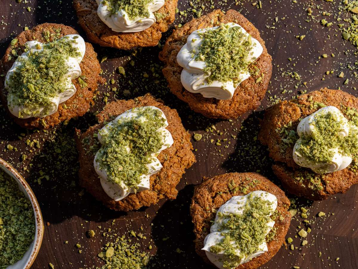The image shows several cookies topped with piped cream and sprinkled with green crumbs, likely matcha or pistachio. They are displayed on a dark wooden surface with some crumbs scattered around, adding a rustic and appetizing presentation.