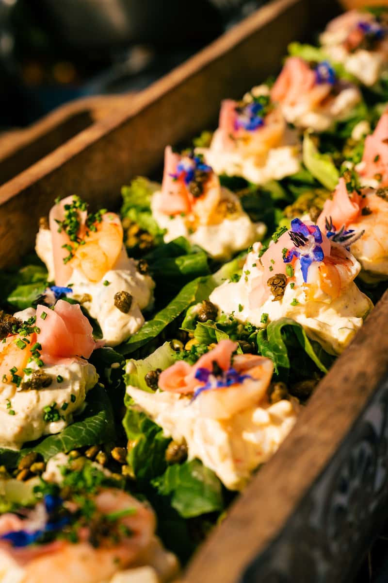 Shrimp salad bites on lettuce leaves, topped with pickled ginger, capers, edible flowers, and fresh herbs in a wooden tray.