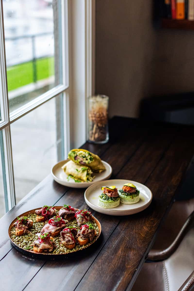 Plated café dishes on a wooden table by the window, featuring a wrap, savory bites, and crostini topped with meat and pickled onions.