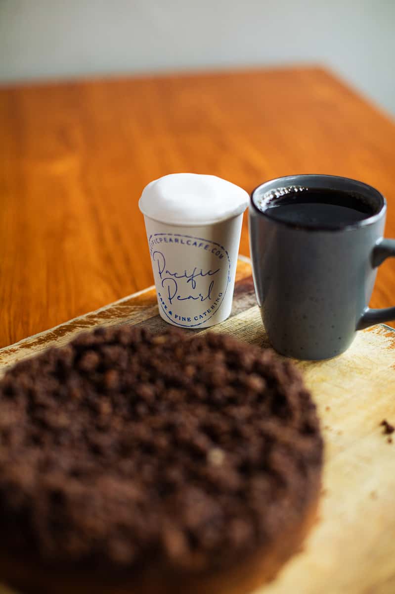 Two cups of coffee, one black and one with foam, sit on a wooden board beside a chocolate crumb cake.