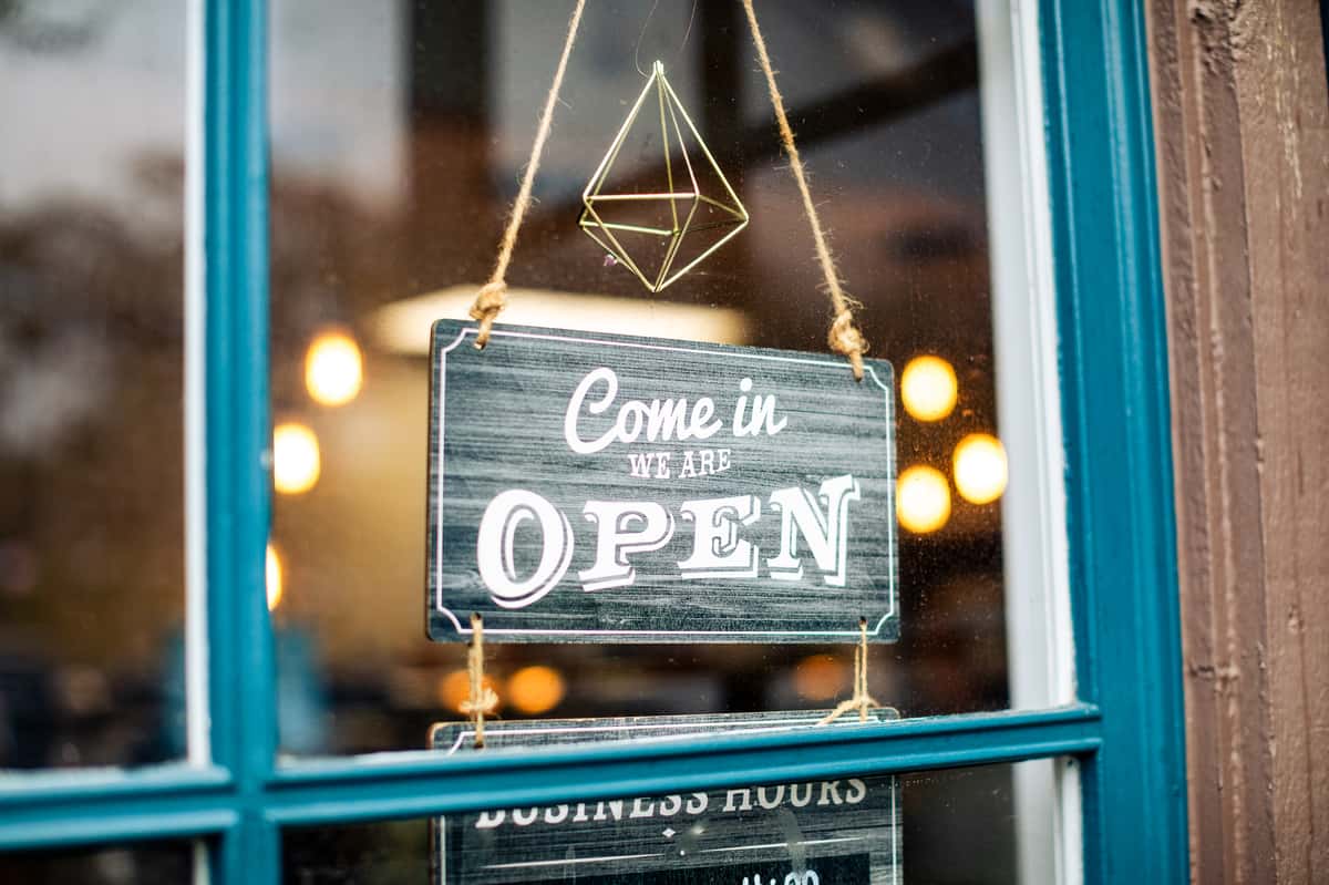 Hanging sign on a glass door that reads "Come in, we are OPEN" with warm lights blurred in the background.
