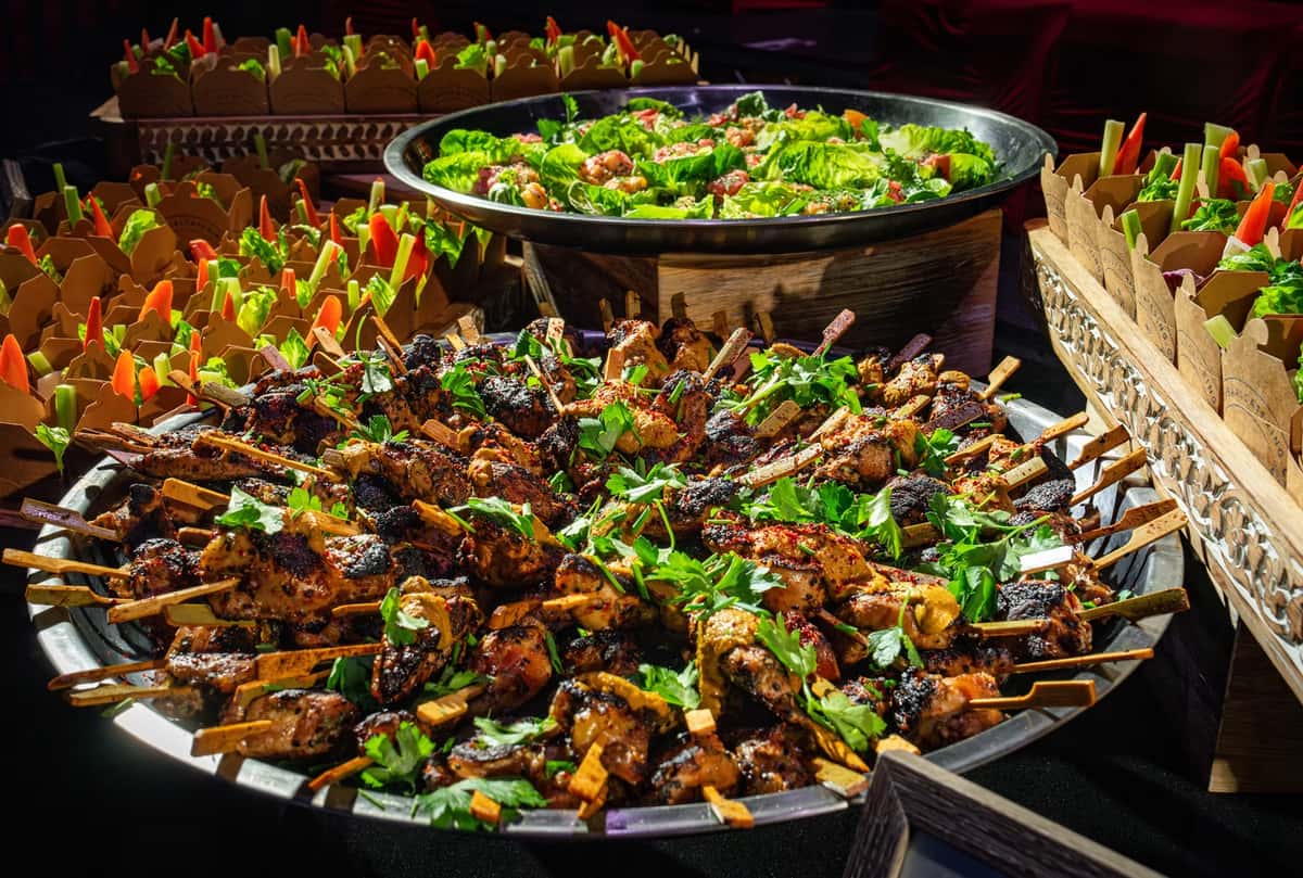 Grilled chicken skewers garnished with herbs, alongside lettuce cups with shrimp and colorful veggie sticks in takeout boxes.