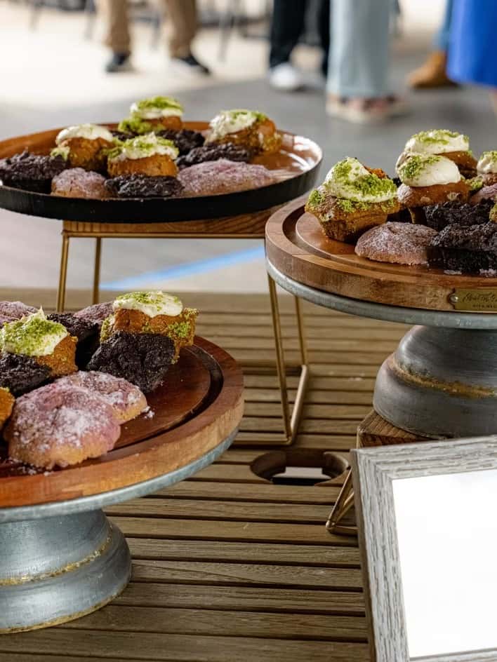 Assorted pastries topped with cream and green crumbles are displayed on wooden stands at an event.
