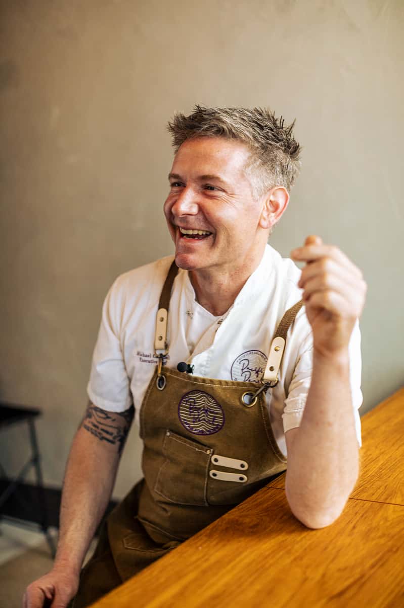 Smiling chef in a brown apron and white uniform sits at a wooden table, appearing cheerful and engaged.