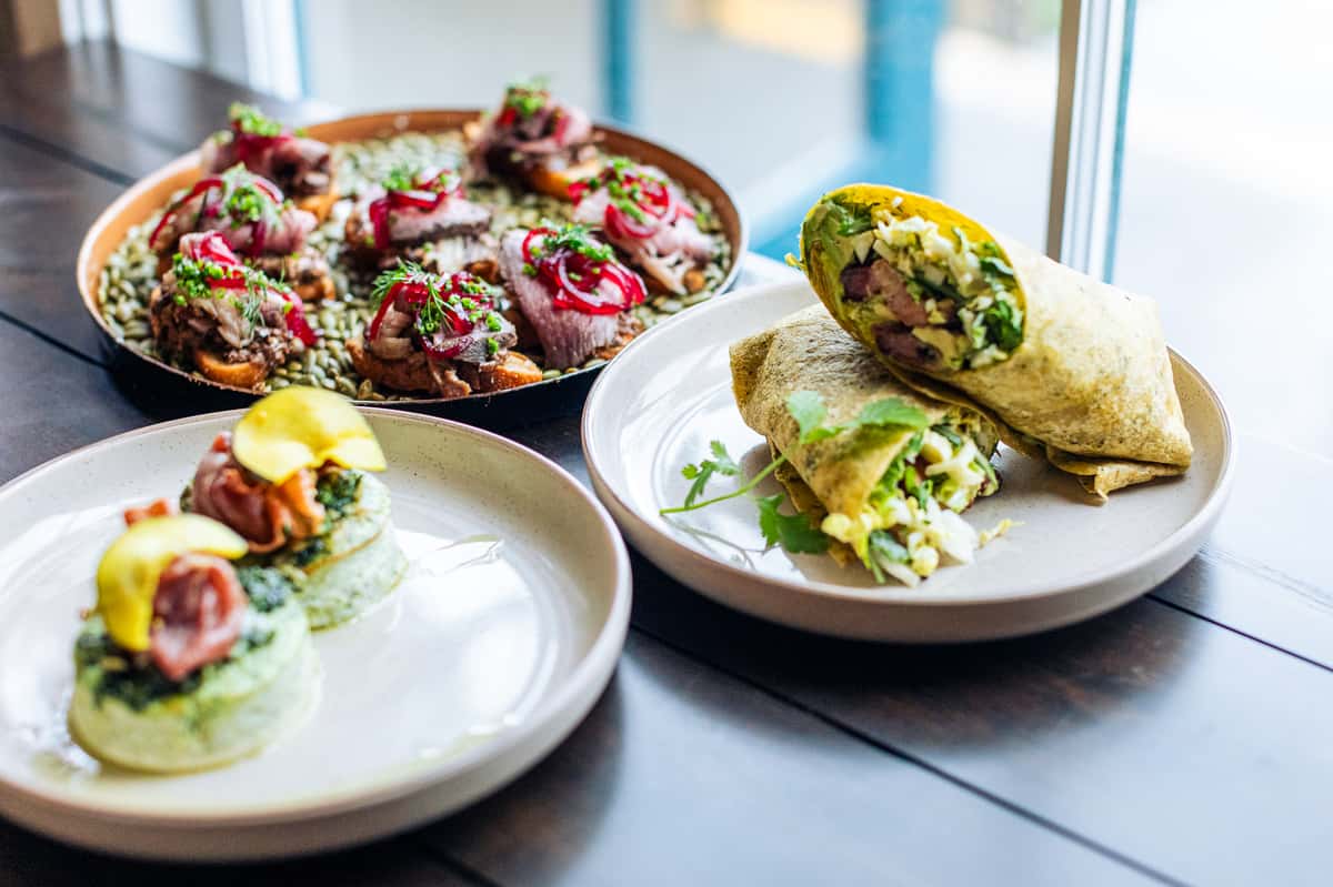 Plates of gourmet dishes with a wrap stuffed with greens, small bites topped with meat and pickled onions, and egg-based entrees.