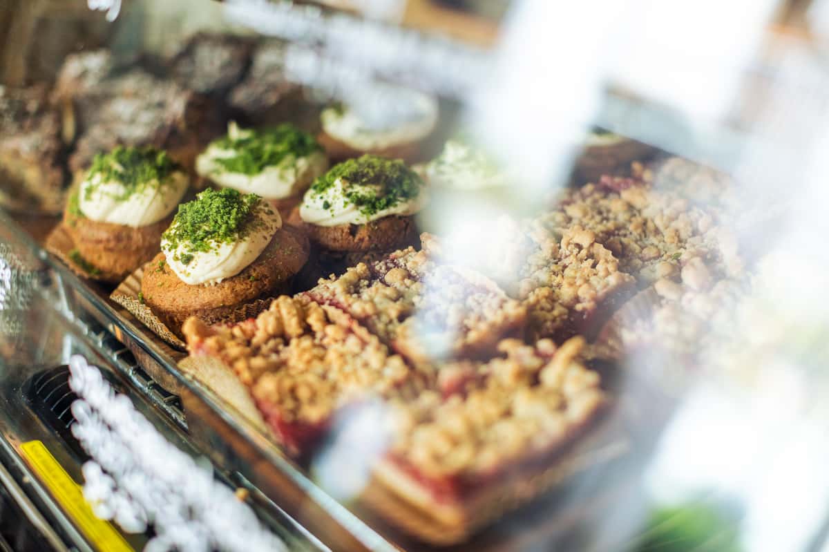 Pastries displayed in a glass case, including frosted muffins topped with green garnish and crumb-topped fruit bars.
