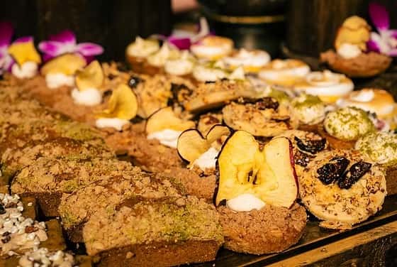Assorted pastries on display, including loaves with crumb topping, tarts with dried fruit, and desserts garnished with apple slices.