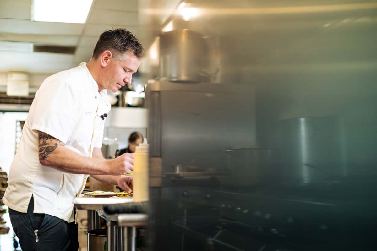 Chef focused on plating a dish in a professional kitchen, with stainless steel equipment and a warm, busy atmosphere.