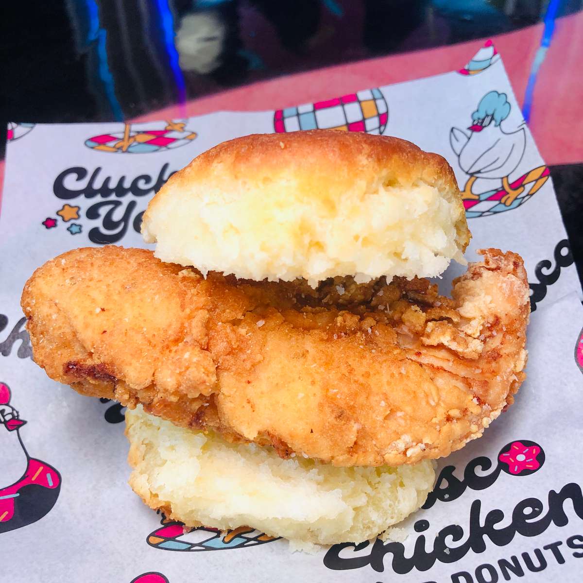 Southern-style fried chicken biscuit sandwich featuring a golden, crispy chicken tender tucked between two fluffy buttermilk biscuits, served on branded paper from a playful chicken and donuts eatery — a bold twist on classic comfort food.