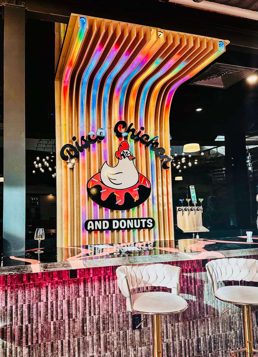 Colorful, retro-inspired interior of Disco Chicken and Donuts featuring a bold neon-lit wall with a cartoon chicken atop a donut, sleek white bar stools, and a glossy counter — a playful, modern twist on a nostalgic diner vibe.