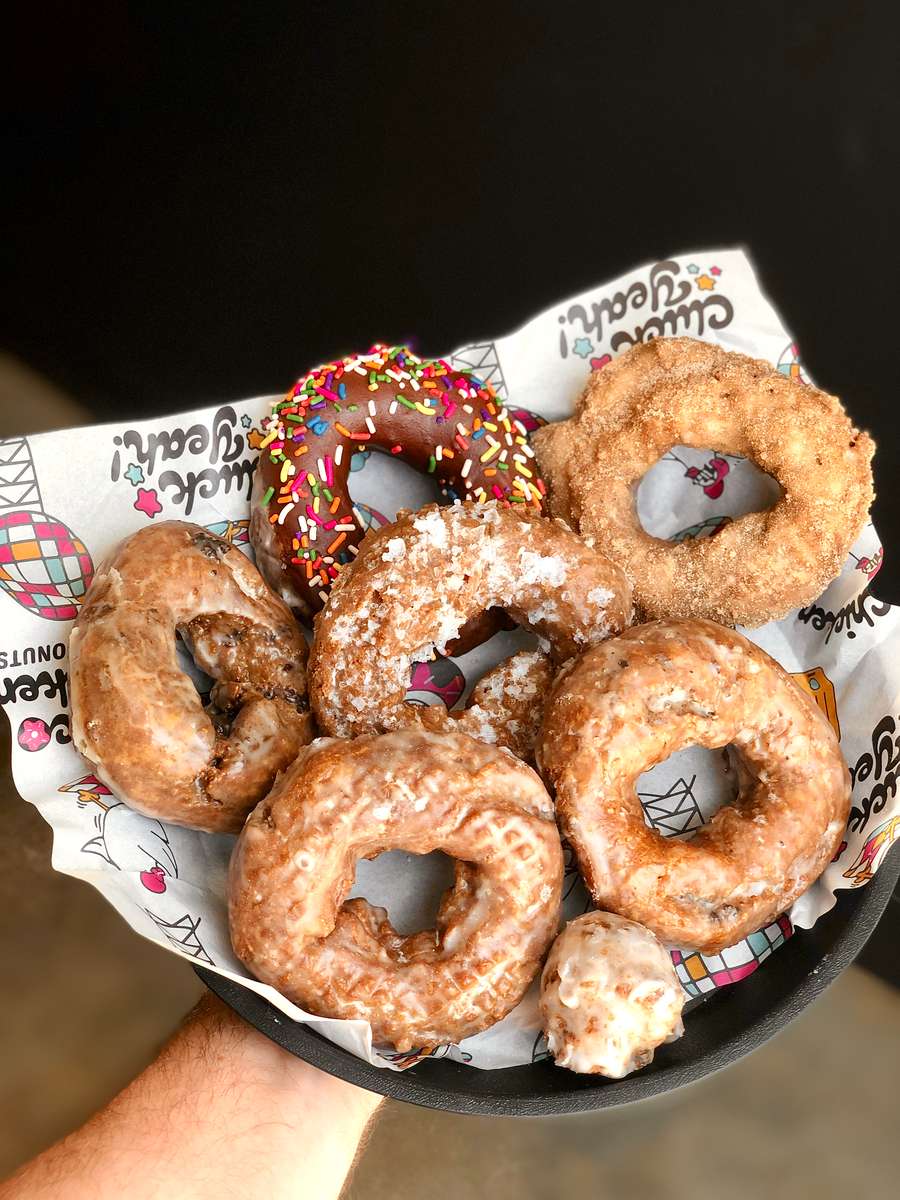 Assorted artisan donuts in a round black basket lined with playful branded paper, featuring old-fashioned glazed, cinnamon sugar, a chocolate frosted donut with rainbow sprinkles, and a glazed donut hole — a fun and flavorful selection from a chicken and donuts restaurant.