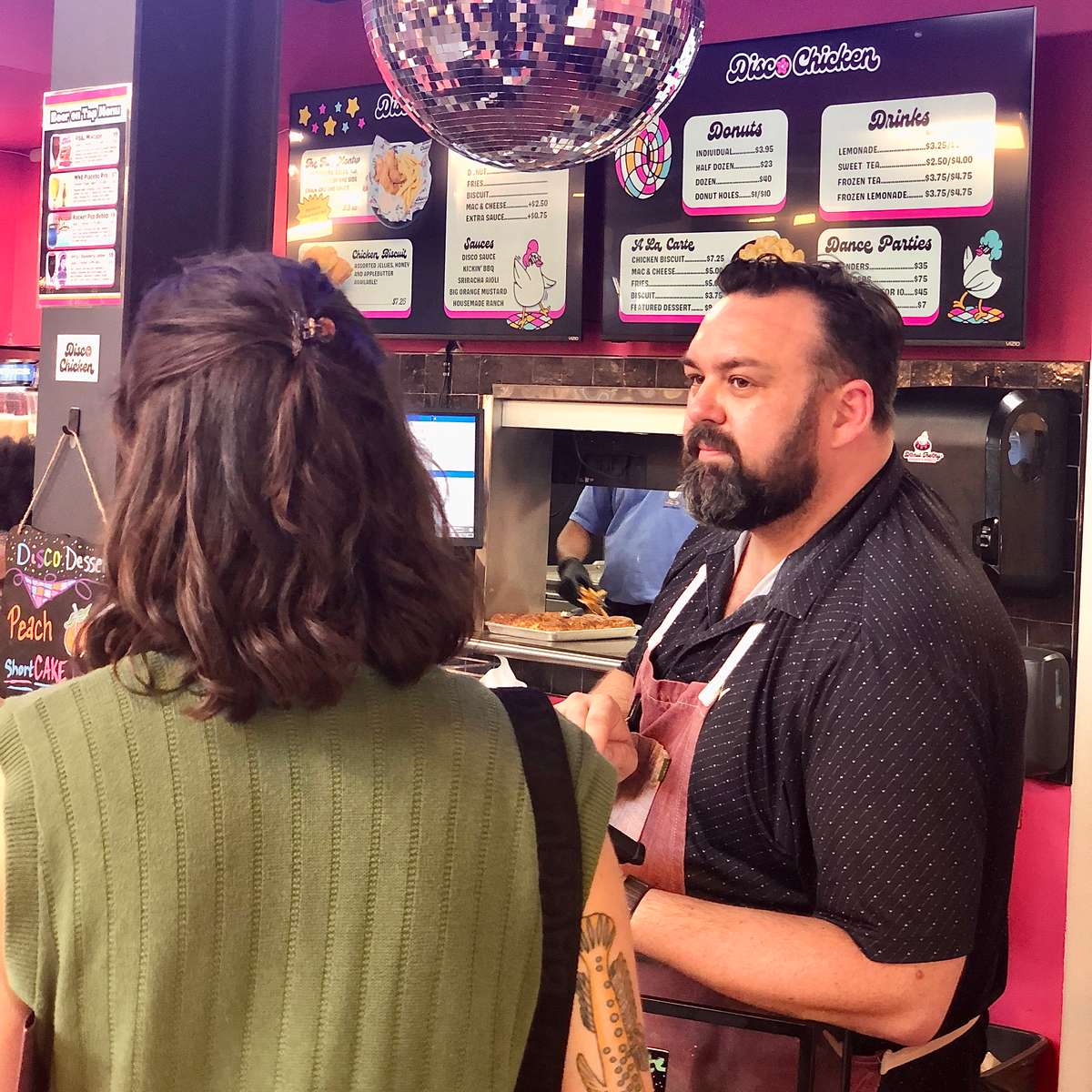 Customer ordering from a staff member at a retro-themed chicken and donuts counter, with a disco ball overhead and colorful menu boards featuring donuts, drinks, and fried chicken dishes — a lively, nostalgic eatery experience.