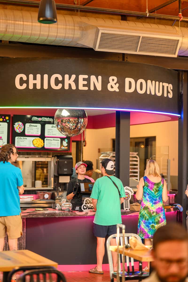 Customers ordering at a vibrant "Chicken & Donuts" counter inside a modern food hall, featuring a neon-lit disco theme with a shiny mirror ball, colorful signage, and a casual, fun atmosphere perfect for fried comfort food lovers.