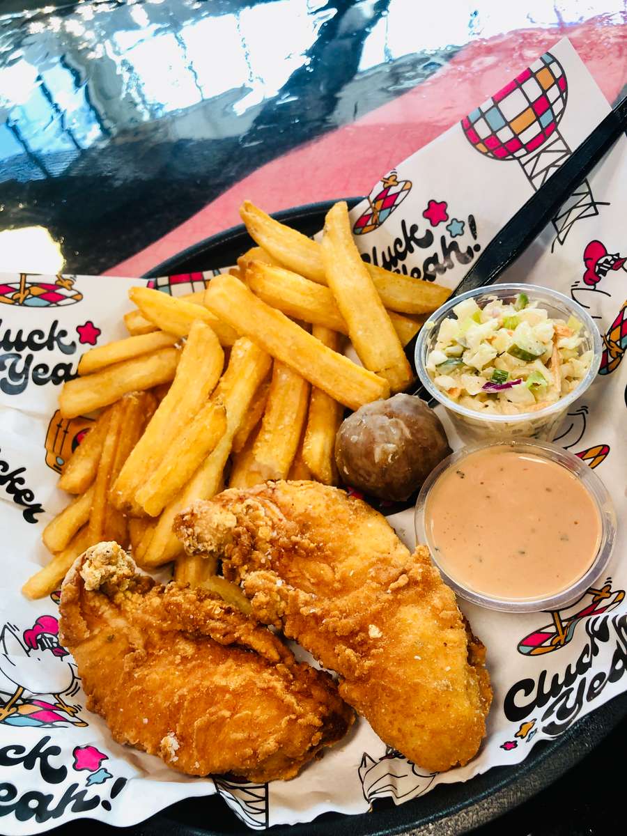 Crispy fried chicken tenders served with thick-cut fries, creamy coleslaw, a glazed donut hole, and a side of zesty dipping sauce, all plated on branded paper with playful "Cluck Yeah!" graphics — a bold and indulgent American comfort food combo.