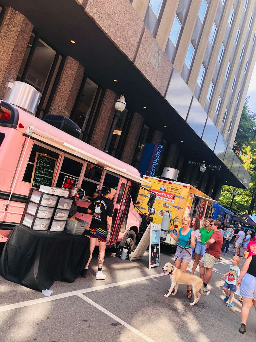 Crowds gathered at a lively downtown food truck event, featuring a retro pink chicken and donuts bus and other colorful food trucks parked along the street, with people and pets enjoying the vibrant outdoor atmosphere.