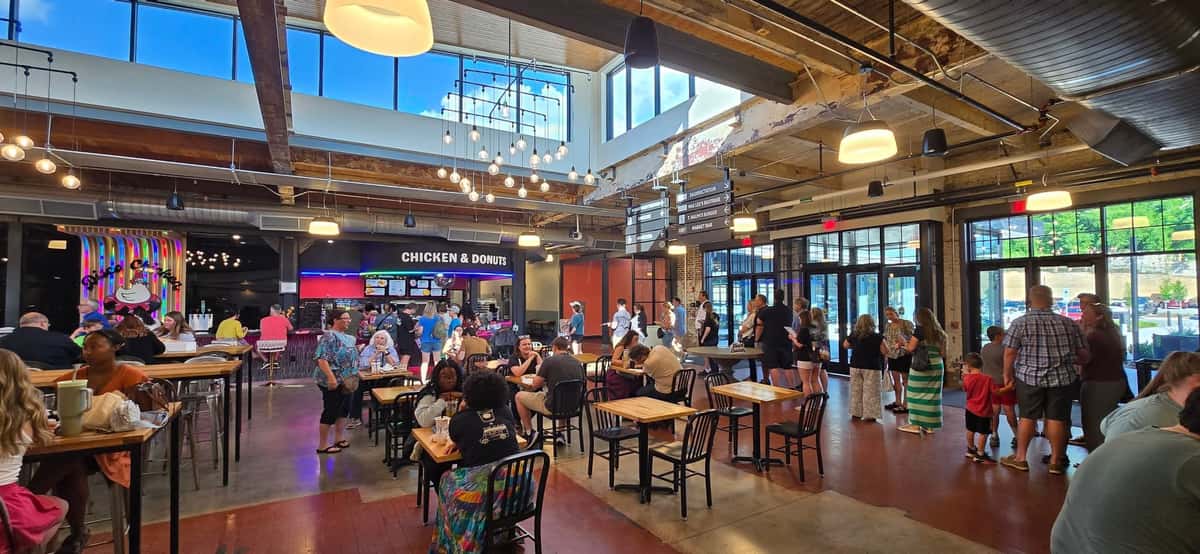 Spacious indoor food hall bustling with diners and visitors, featuring high industrial ceilings, large windows, and a modern chicken and donuts counter at the back — a trendy, family-friendly dining spot with casual seating and vibrant decor.