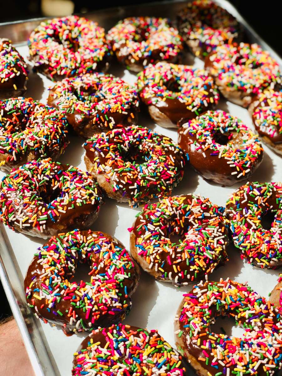 Tray of freshly made chocolate glazed donuts topped with colorful rainbow sprinkles, displayed in rows under natural light — a vibrant and playful sweet treat from a specialty donut shop.