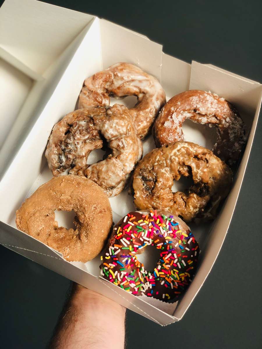 Assorted gourmet donuts in a white takeout box, featuring glazed old-fashioned donuts, a cinnamon sugar donut, and a chocolate-frosted donut topped with colorful rainbow sprinkles — perfect for a classic bakery breakfast or sweet treat.