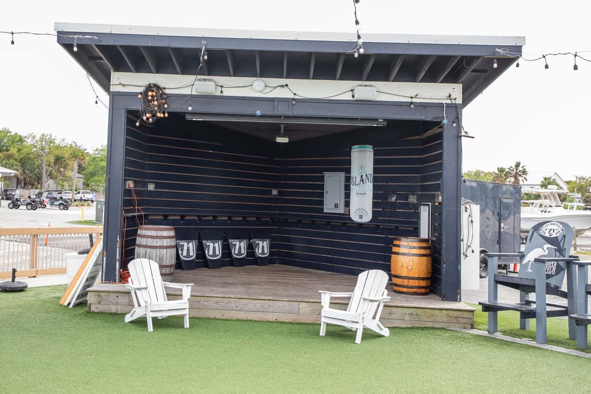 open-air coastal bar stage with wooden barrels, Adirondack chairs, and string lights on a grassy patio
