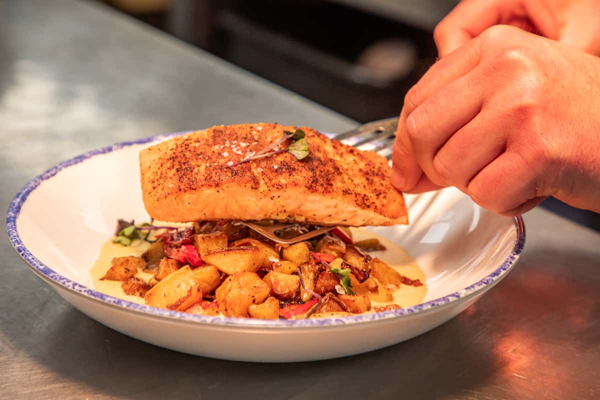 chef finishing a plated salmon entrée with vegetables in a professional kitchen