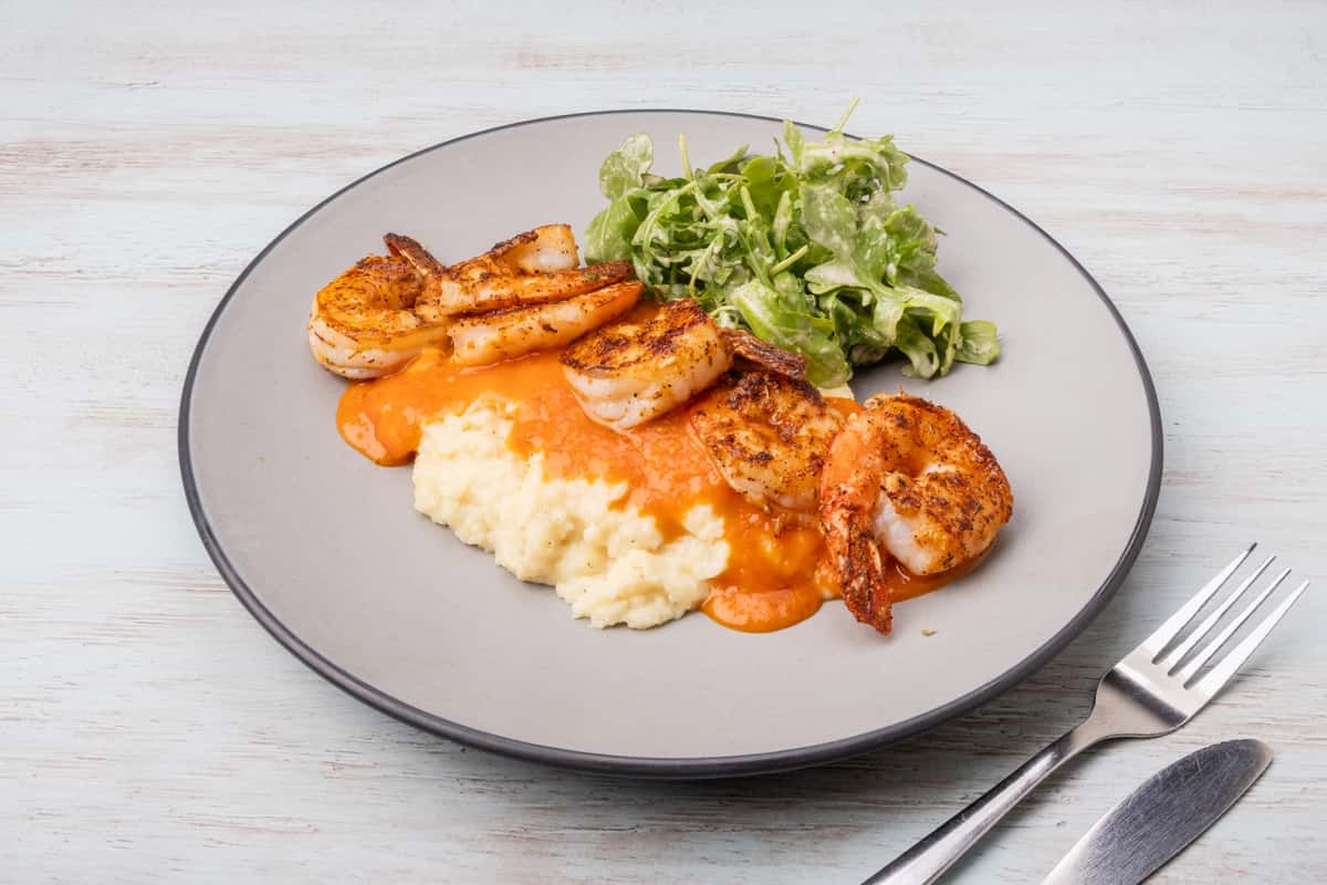 Grilled shrimp and creamy grits topped with a spicy sauce, served with a side salad on a gray plate at a restaurant