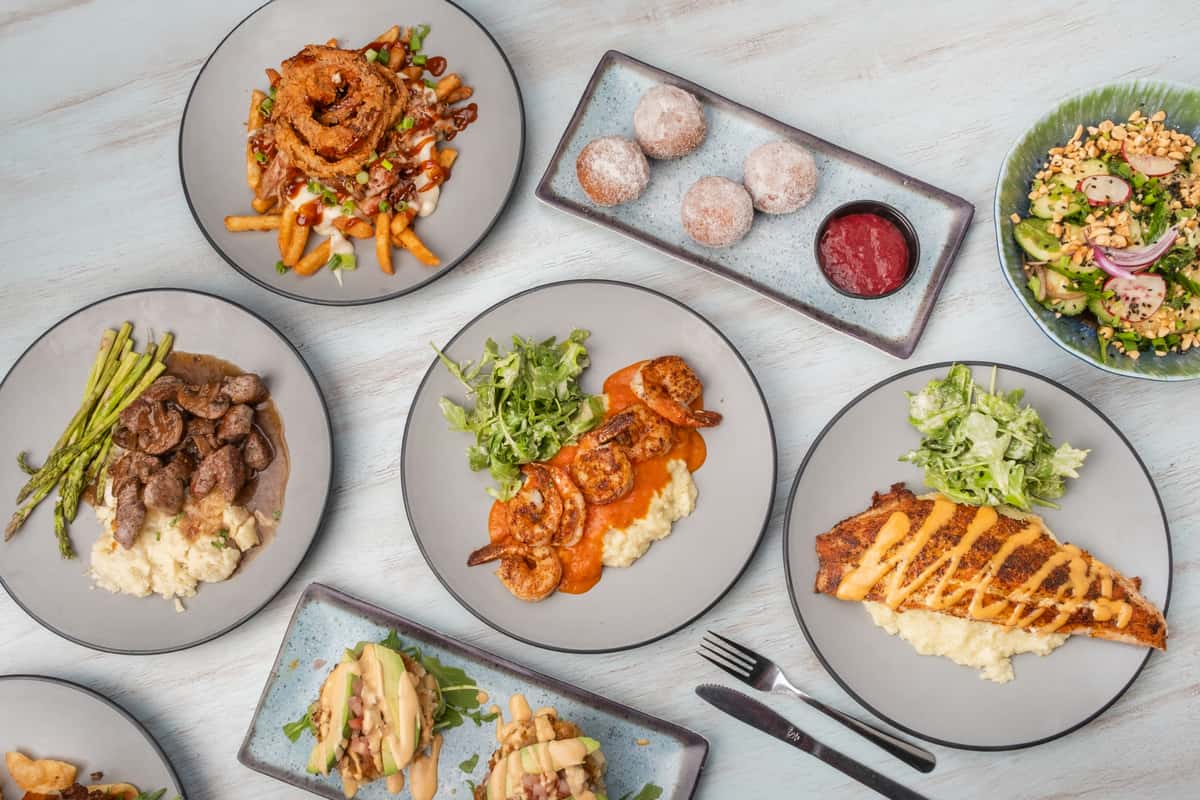 Assorted restaurant dishes displayed together, including an Asian-style noodle salad with shrimp, sliders with dipping sauce, steak tips with mashed potatoes, and sandwiches on gray plates