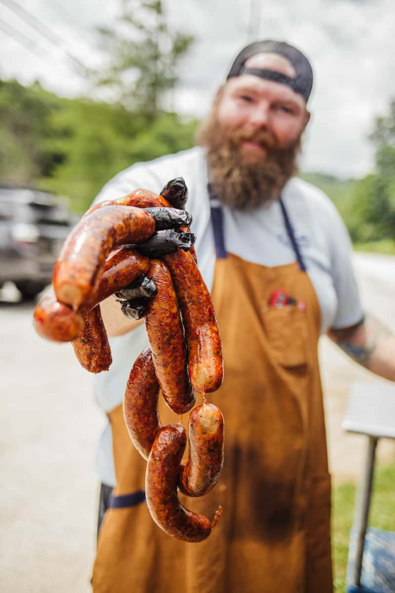 A bearded man in a brown apron holds out a handful of sausages. He is probably a butcher or cook proud of the meats offered by the restaurant.