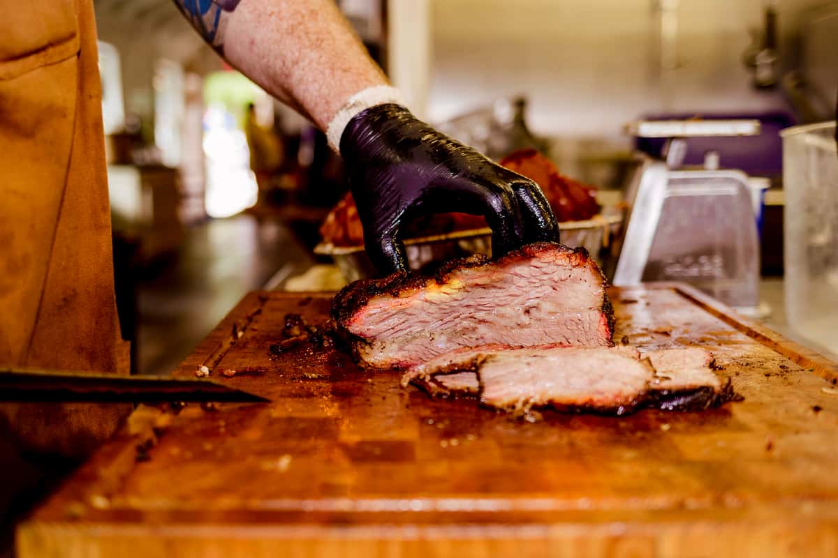 Close-up of freshly smoked brisket being sliced on a wooden cutting board. The meat has a smoky bark and pink smoke ring.