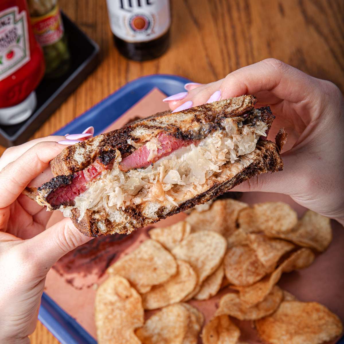 Close up of a Reuben sandwich held in two hands, alongside potato chips. The sandwich is made with rye bread, corned beef, sauerkraut, and Swiss cheese. This image is used as a header image on a BBQ restaurant website.
