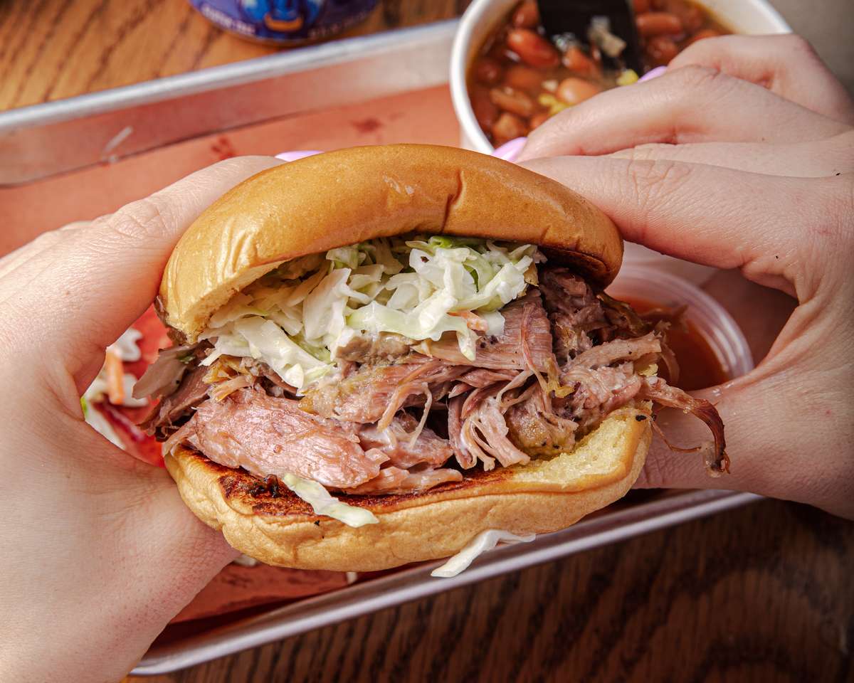 A pulled pork sandwich with coleslaw. The sandwich is being held in two hands, and there is a side of beans in the background.
