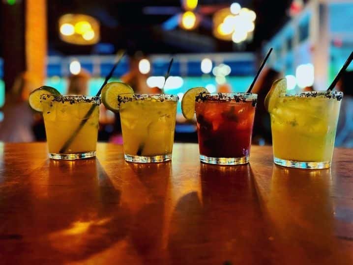Four vibrant margaritas garnished with lime wheels and black salt rims, lined up on a wooden bar top in a dimly lit, modern restaurant with a lively ambiance.