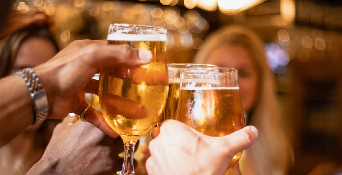Close-up of friends clinking glasses of golden draft beer in a warmly lit bar, celebrating with a toast amid a festive, blurred background.