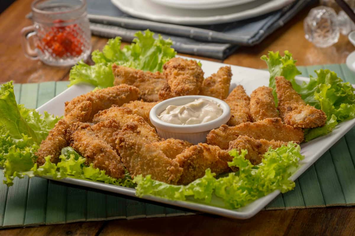 Plated crispy chicken fingers arranged on a bed of leafy greens, paired with a ramekin of creamy dipping sauce in a rustic dining setting.