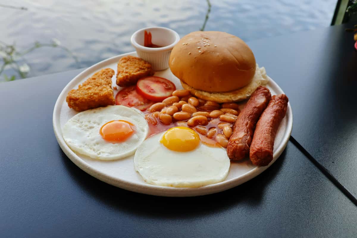 Full breakfast plate with two fried eggs, sausages, baked beans, hash browns, sliced tomatoes, a bread roll, and ketchup.