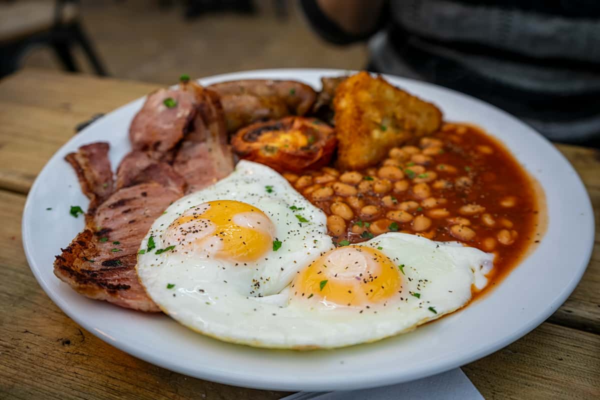 Two fried eggs with bacon, sausages, grilled tomato, hash brown, and baked beans on a white plate.