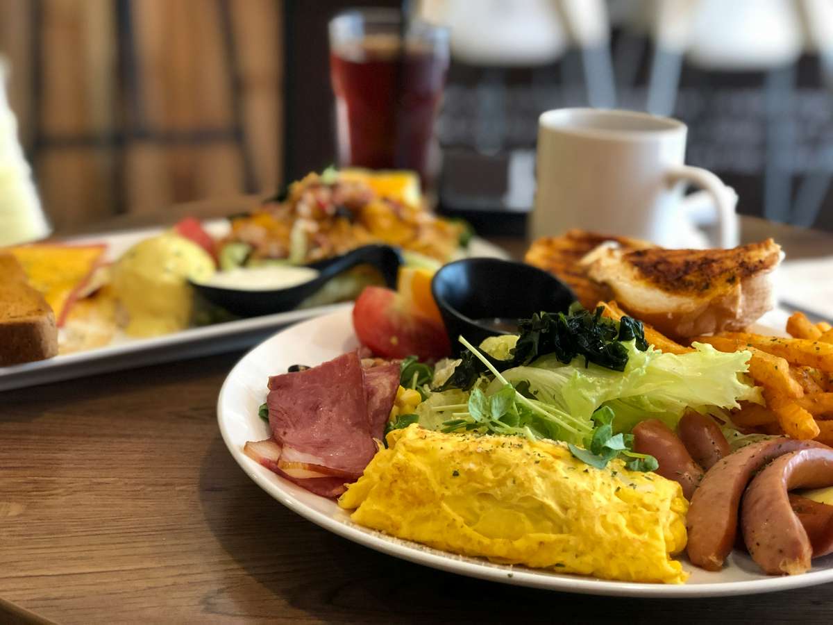 Café breakfast plate with an omelet, sausages, bacon, salad, fries, and toast, with another meal and drinks blurred in the background.